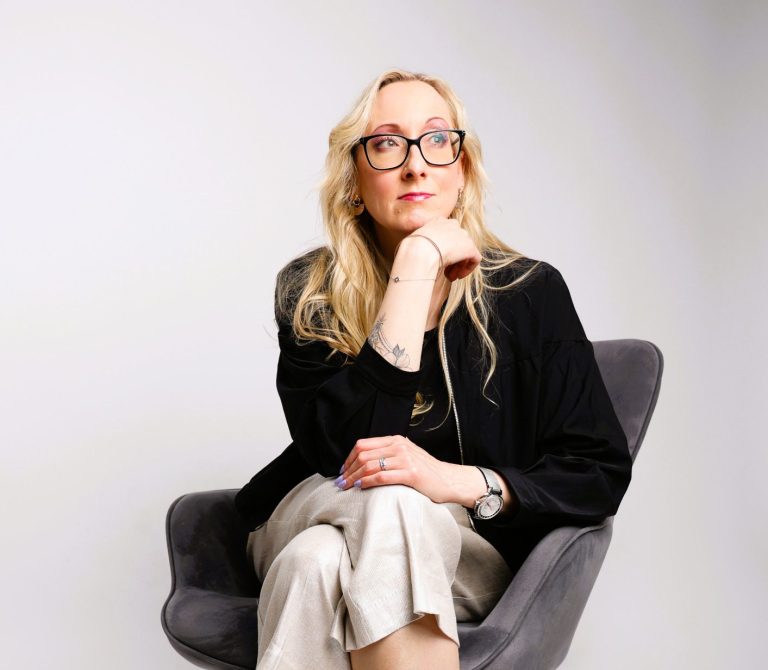 Junge Frau mit Brille, lächelnd, in einem hellen Raum, trägt ein Outfit aus Blazer und Jeans.