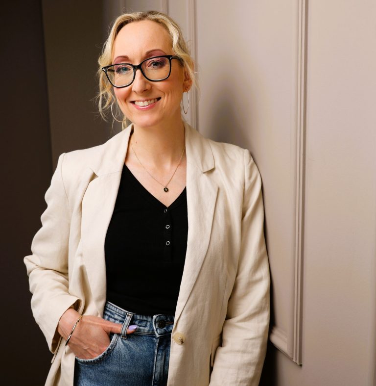 Junge Frau mit Brille, lächelnd, in einem hellen Raum, trägt ein Outfit aus Blazer und Jeans.